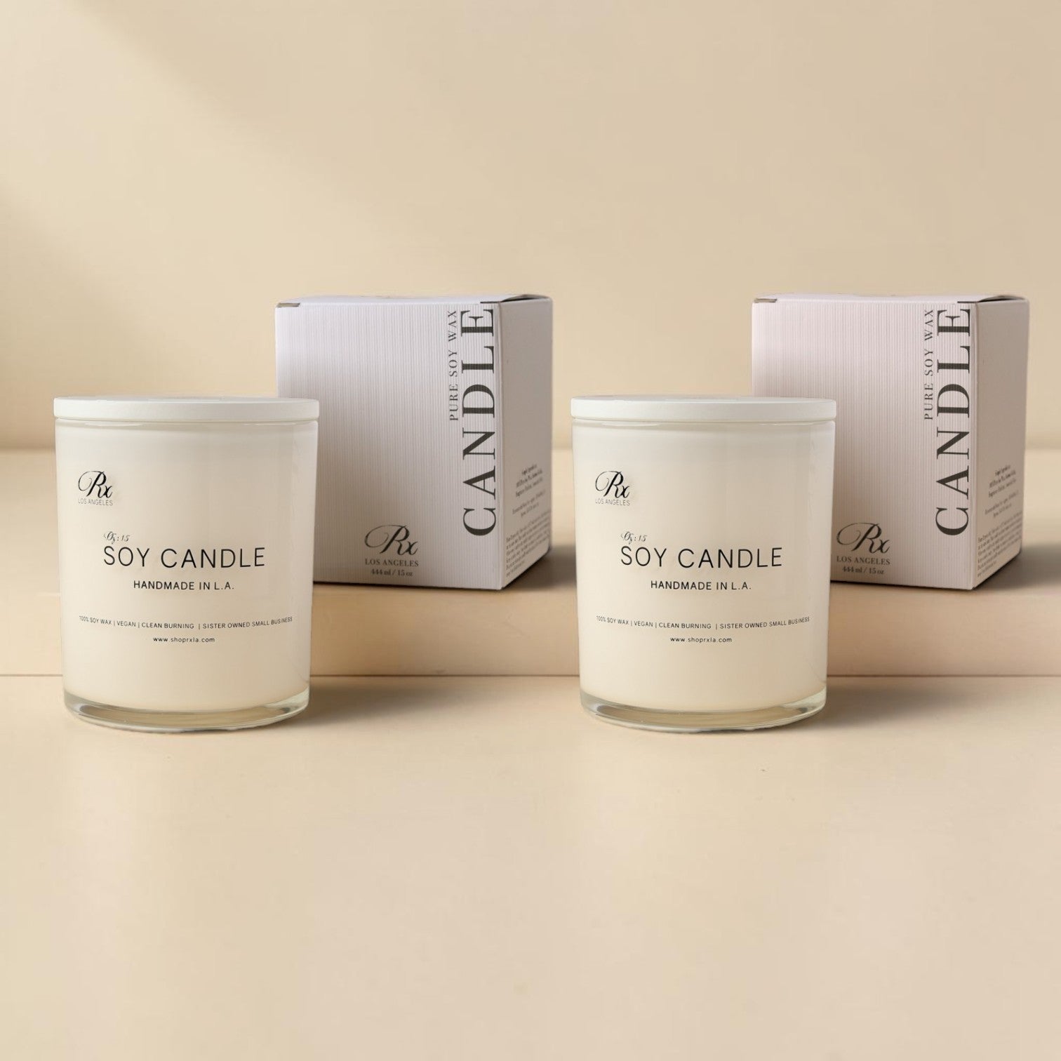 Two white soy candles in  glass jars with white lids are shown in front of their matching white packaging boxes. The labels read Soy Candle Handmade in LA'.
