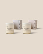 Two soy candles in clear glass jars with white lids and labels, shown in front of their matching pale pink and white boxes on a solid beige background.