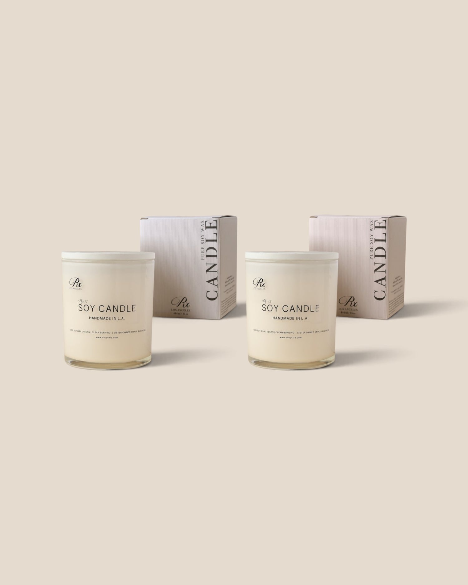 Two soy candles in clear glass jars with white lids and labels, shown in front of their matching pale pink and white boxes on a solid beige background.