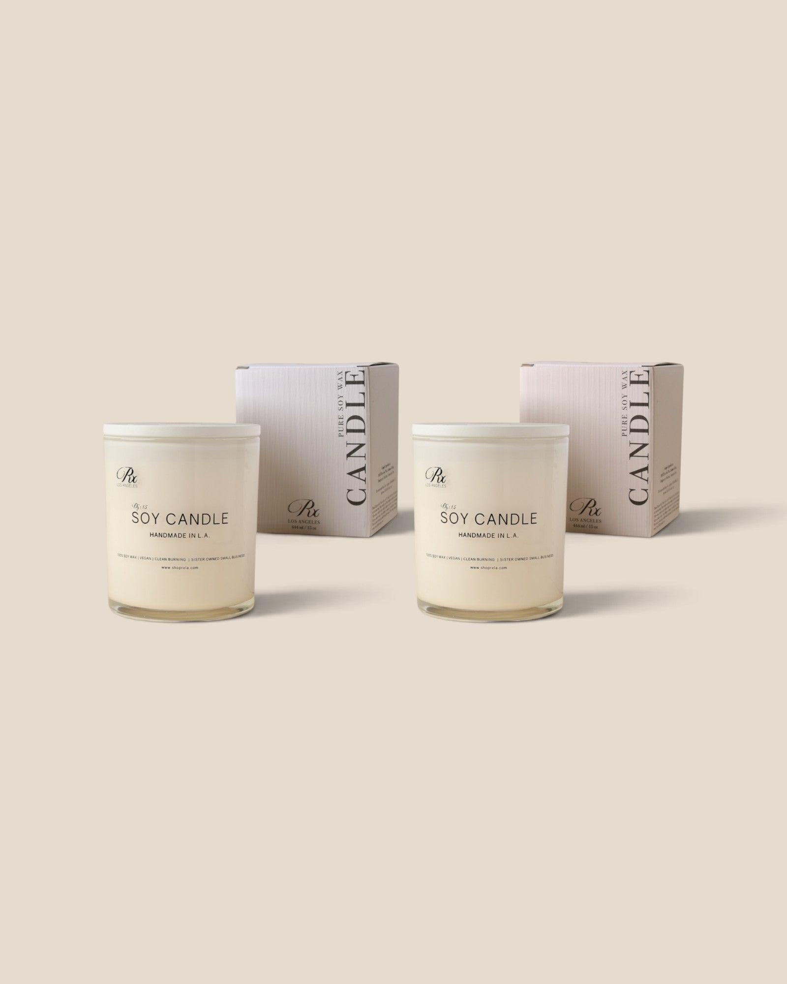 Two soy candles in clear glass jars with white lids and labels, shown in front of their matching pale pink and white boxes on a solid beige background.