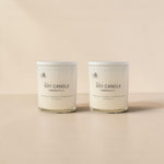 Two white soy candles in glass jars with white lids, standing side by side on a warm beige surface against a matching beige background. The labels read 'The Artisan' and 'Soy Candle Handmade in LA'.