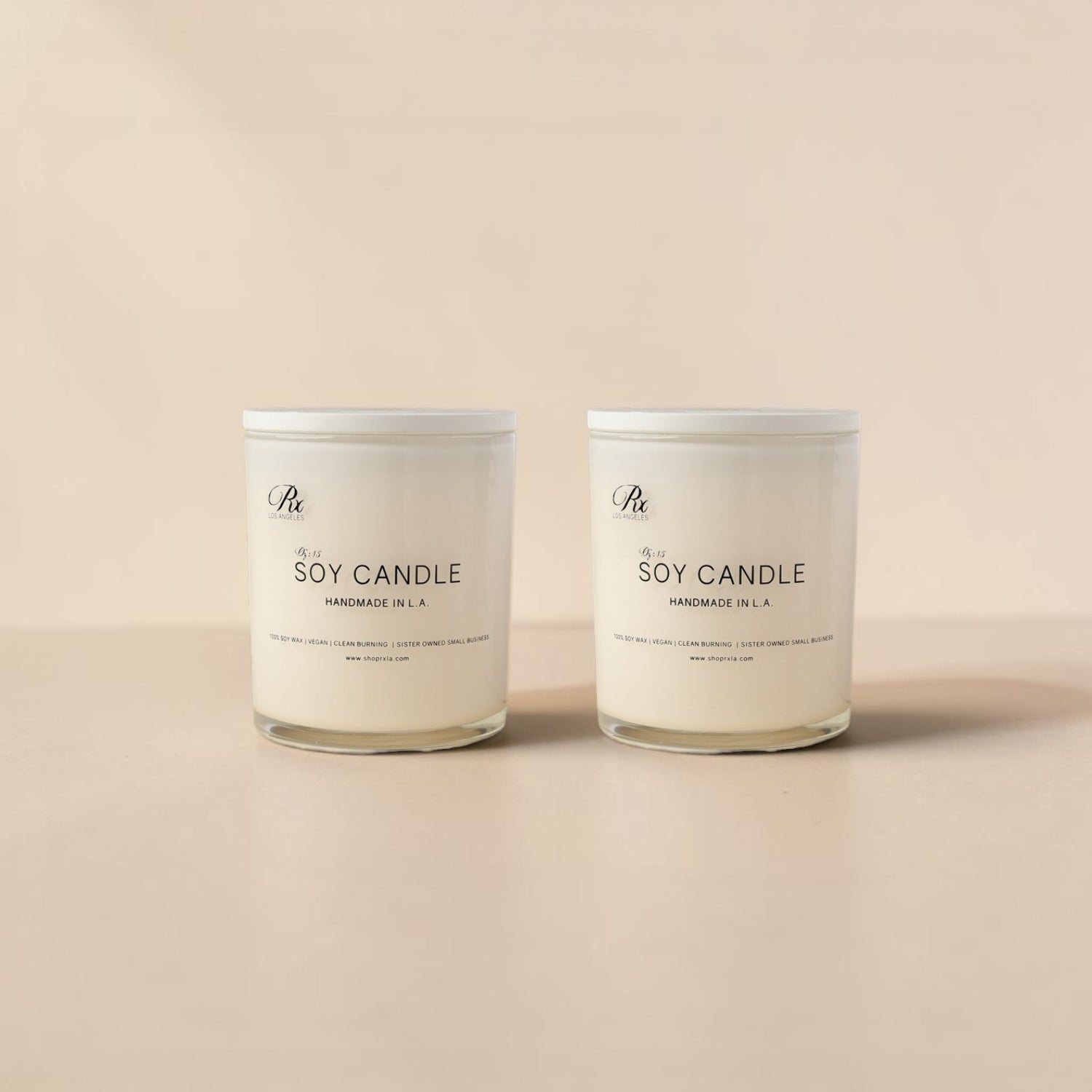 Two white soy candles in glass jars with white lids, standing side by side on a warm beige surface against a matching beige background. The labels read 'The Artisan' and 'Soy Candle Handmade in LA'.
