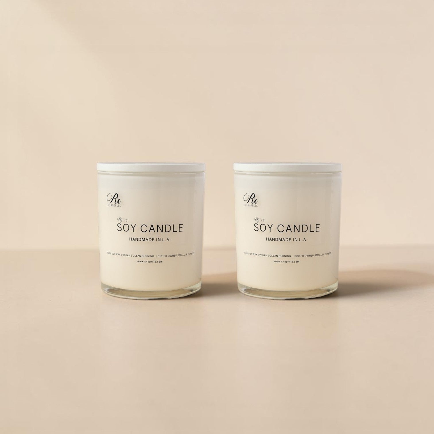 Two white soy candles in glass jars with white lids, standing side by side on a warm beige surface against a matching beige background. The labels read 'The Artisan' and 'Soy Candle Handmade in LA'.
