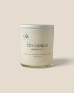 A soy candle in a clear glass jar with a white lid, featuring minimalist black text on the front label, set against a solid beige background.