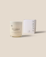 A soy candle in a clear glass jar with cream-colored wax and a white lid, shown next to its white packaging box on a solid beige background.