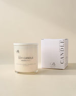 A clear glass jar candle with white wax sits beside a white product box on a warm cream background. The label reads 'The Artisan Candle' and 'SOY CANDLE HANDMADE IN A.