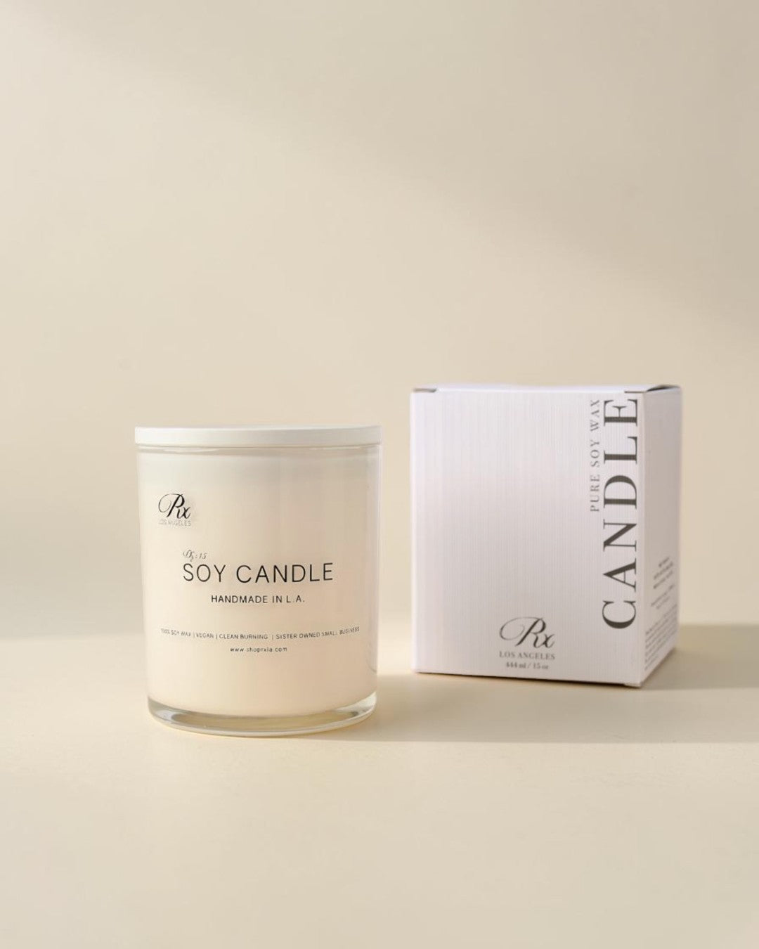 A clear glass jar candle with white wax sits beside a white product box on a warm cream background. The label reads 'The Artisan Candle' and 'SOY CANDLE HANDMADE IN A.