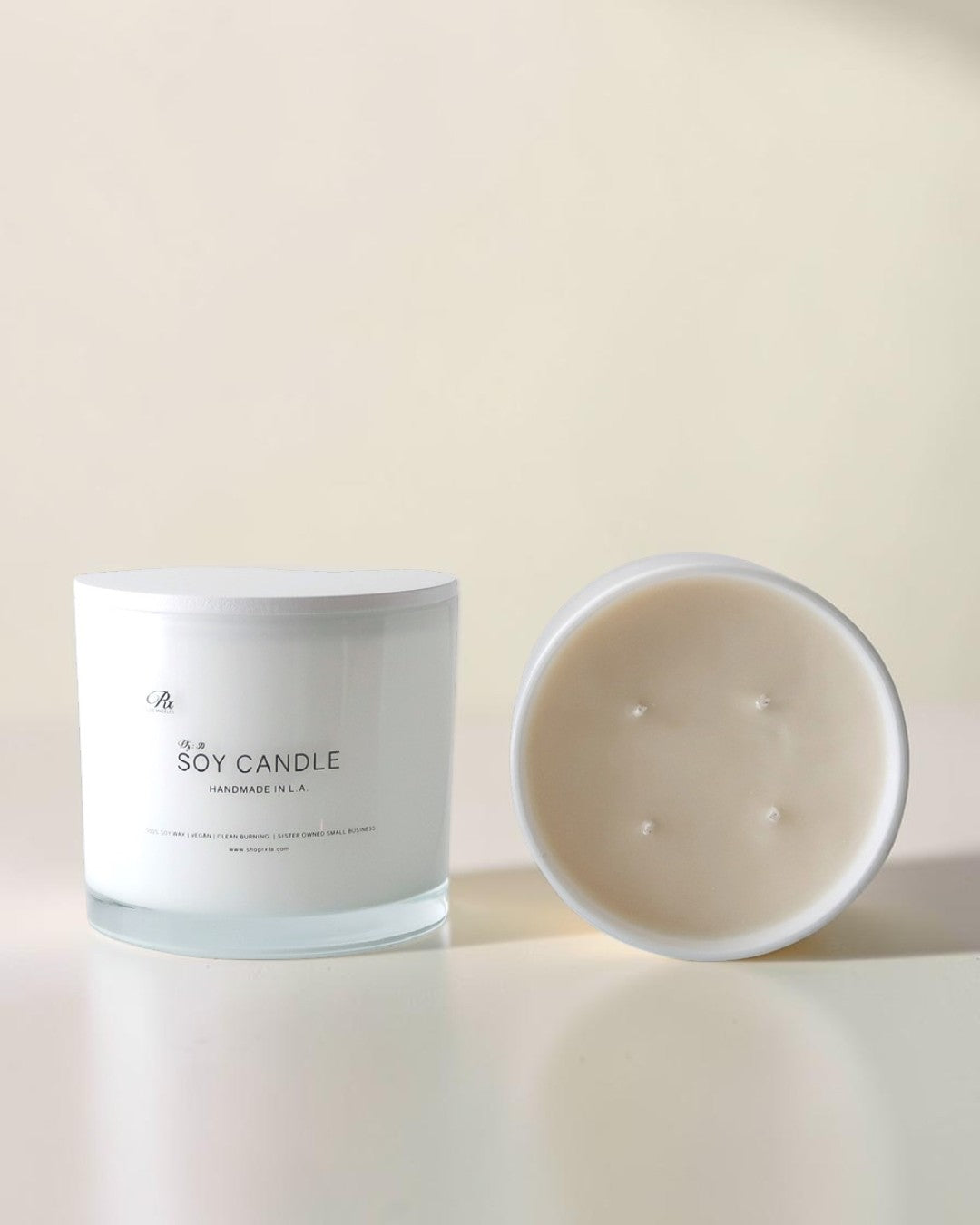 A soy candle in a frosted glass jar with its lid placed beside it on a clean, light beige surface. The label reads 'SOY CANDLE' with a small logo above it.