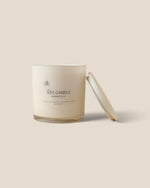 A minimalist soy candle in a clear glass jar with cream-colored wax and a matching lid leaning against it, set against a solid beige background.