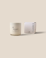 A white soy candle in a clear glass jar sits next to its white packaging box on a solid beige background. The minimalist label and box display elegant black typography.