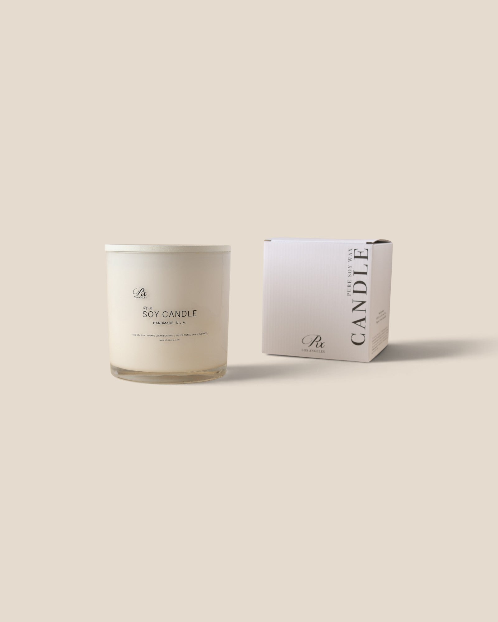 A white soy candle in a clear glass jar sits next to its white packaging box on a solid beige background. The minimalist label and box display elegant black typography.