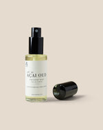 A clear glass spray bottle of Açaí Oud body spray with a black pump and a white label, shown next to its detached black cap on a solid beige background.