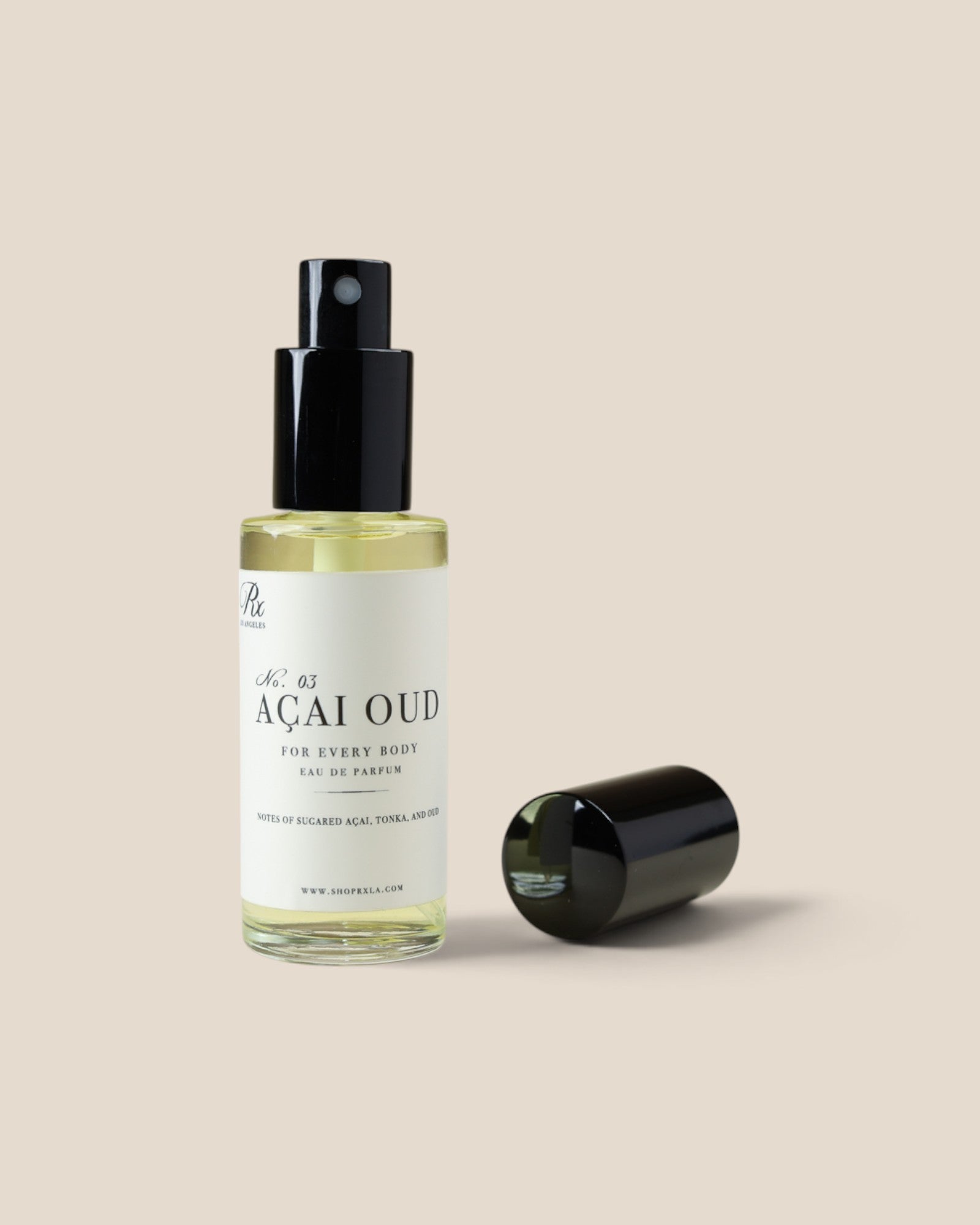 A clear glass spray bottle of Açaí Oud body spray with a black pump and a white label, shown next to its detached black cap on a solid beige background.
