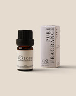 A small amber glass bottle of Acai Oud Pure Fragrance Oil with a black cap stands next to its white striped box on a solid beige background.