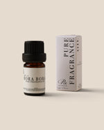 A small amber glass bottle of pure bora bora fragrance oil with a black cap stands next to its white ribbed packaging box on a warm beige background.