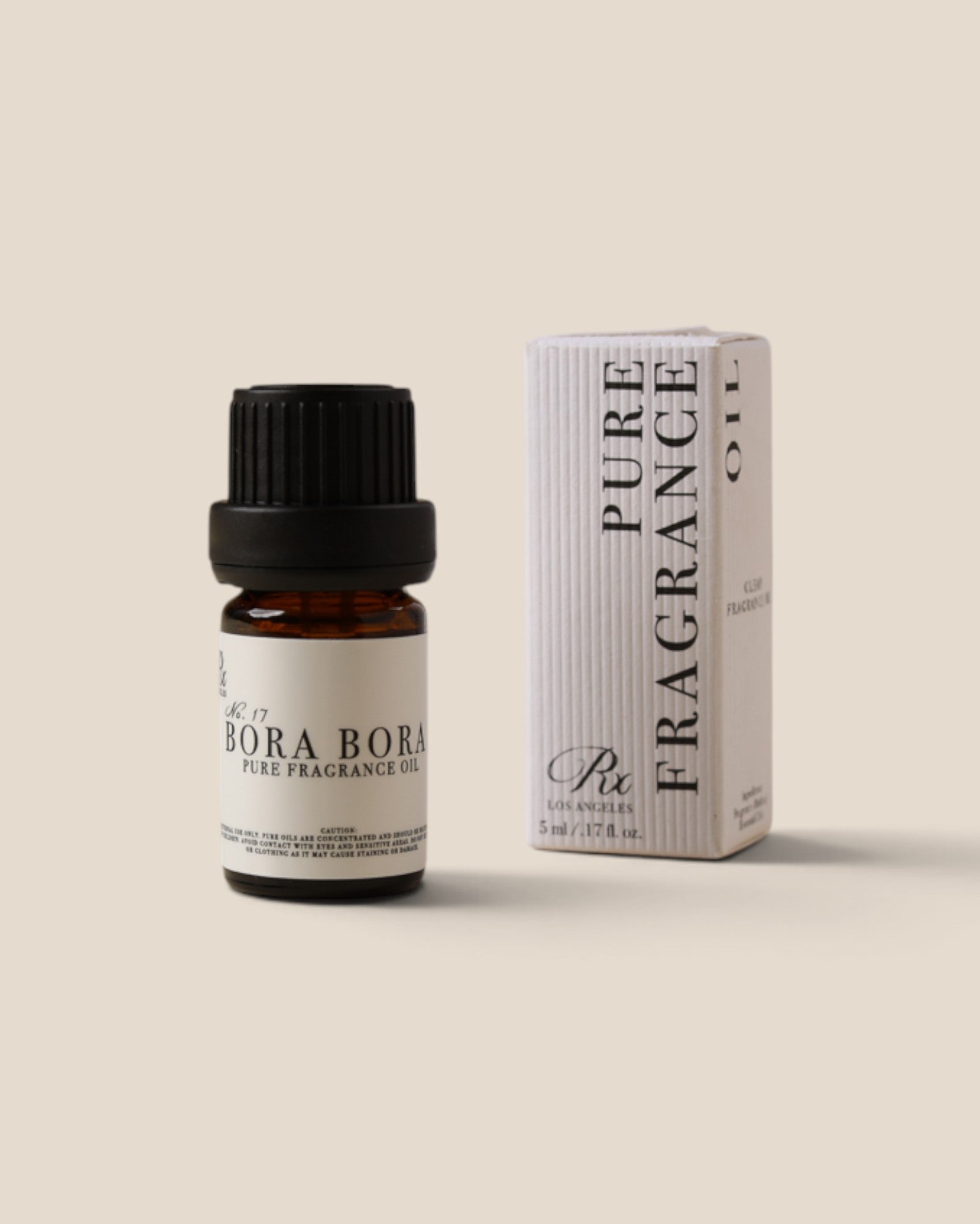 A small amber glass bottle of pure bora bora fragrance oil with a black cap stands next to its white ribbed packaging box on a warm beige background.