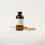 An amber glass reed diffuser bottle with a black cap and a white label reading 'BORA BORA' and 'LUXE DIFFUSER', shown with several natural reed sticks on a clean white background.
