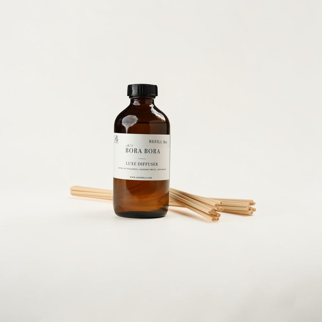 An amber glass reed diffuser bottle with a black cap and a white label reading 'BORA BORA' and 'LUXE DIFFUSER', shown with several natural reed sticks on a clean white background.