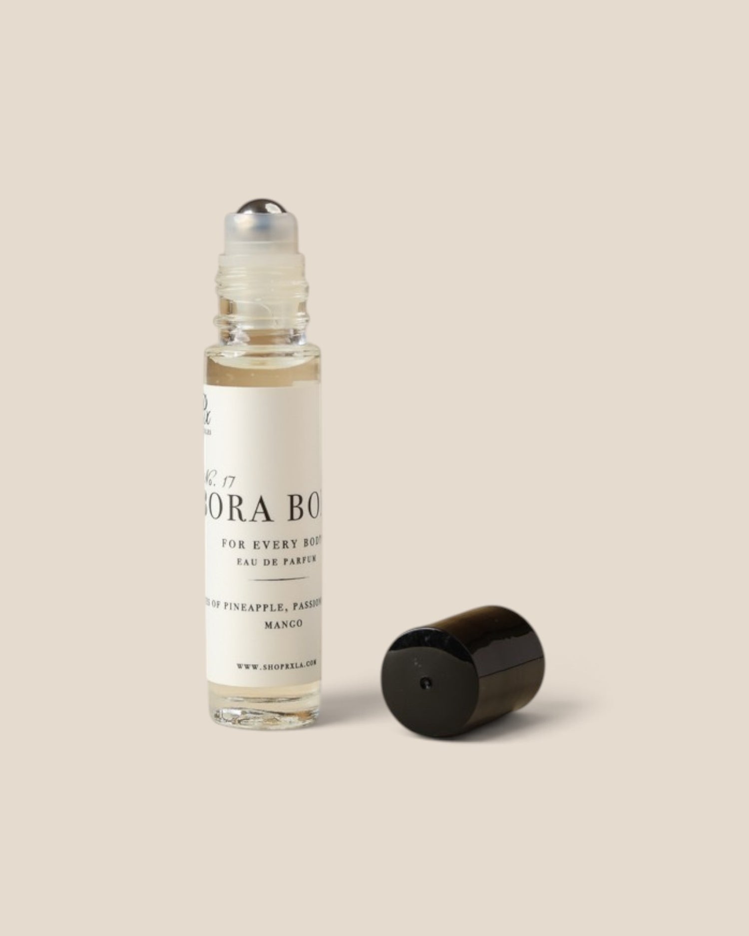 A clear glass rollerball perfume bottle with a white label and black text, shown next to its detached black cap on a solid beige background. 