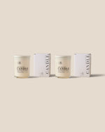 Two glass jar candles with cream-colored wax and white lids, displayed alongside their matching white packaging boxes on a solid beige background.