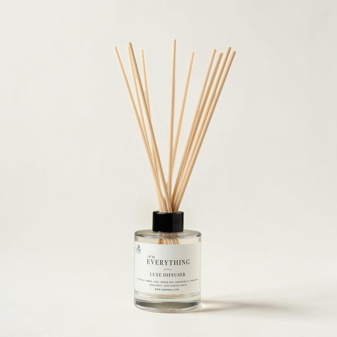 A reed diffuser bottle with a clear glass base, white label, black cap, and natural wood reeds fanning upward, photographed on a clean off-white background.