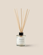 A reed diffuser bottle with a clear glass container, white liquid, a black cap, and diffuser reeds, labeled 'The Fresh Fig Local Diffuser', on a solid beige background.