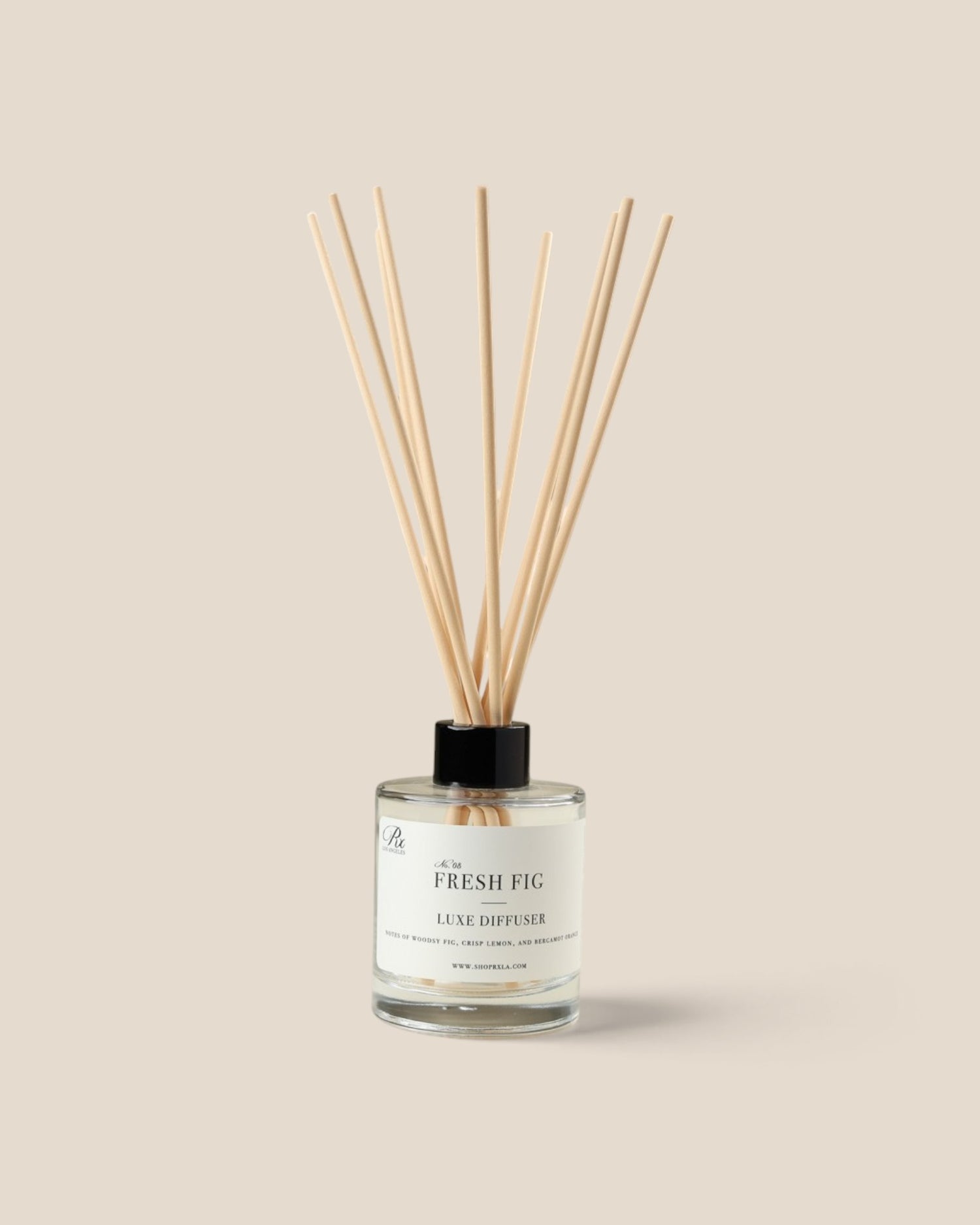 A reed diffuser bottle with a clear glass container, white liquid, a black cap, and diffuser reeds, labeled 'The Fresh Fig Local Diffuser', on a solid beige background.