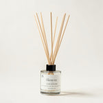 A reed diffuser bottle with a clear glass container, white label, black cap, and natural wood reeds, photographed on a clean off-white background.