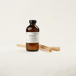 An amber glass bottle of 'The Fresh Fig' Luxe Diffuser refill with a black cap and white label, shown with several natural bamboo diffuser sticks on a clean off-white background.
