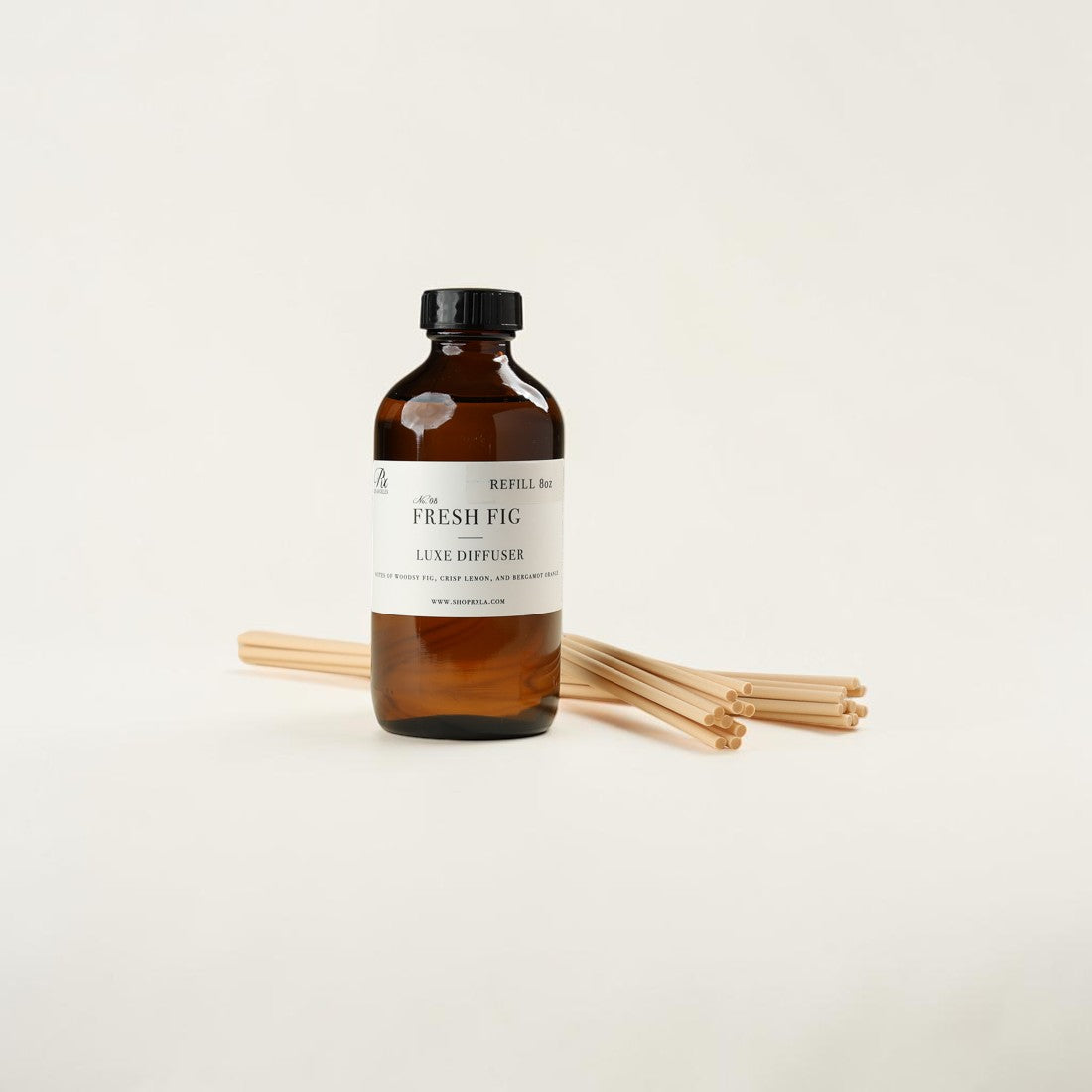 An amber glass bottle of 'The Fresh Fig' Luxe Diffuser refill with a black cap and white label, shown with several natural bamboo diffuser sticks on a clean off-white background.