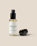 A clear glass spray bottle labeledGARDENIA' with a black pump and a separate black cap placed beside it on a warm beige background.