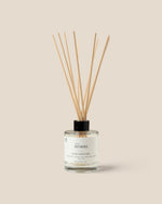 A reed diffuser bottle with a clear glass container, white label, black cap, and fiber reeds, centered on a warm beige background. The label reads 'Homme.