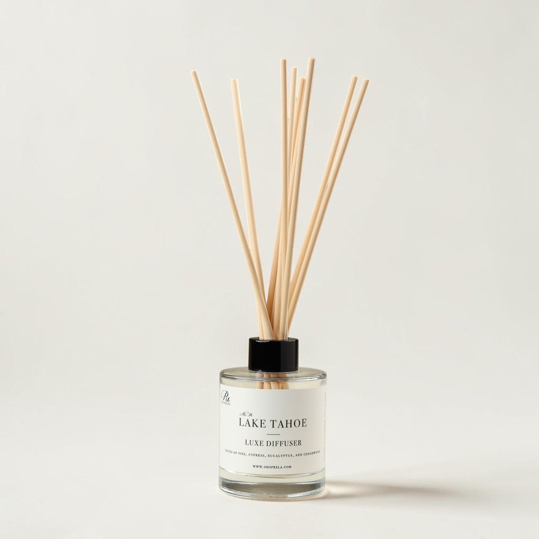 A reed diffuser bottle with a clear glass body, white label reading 'at the LAKE TAHOE LUXE DIFFUSER,' black cap, and natural wood reeds, centered on a light warm gray background.