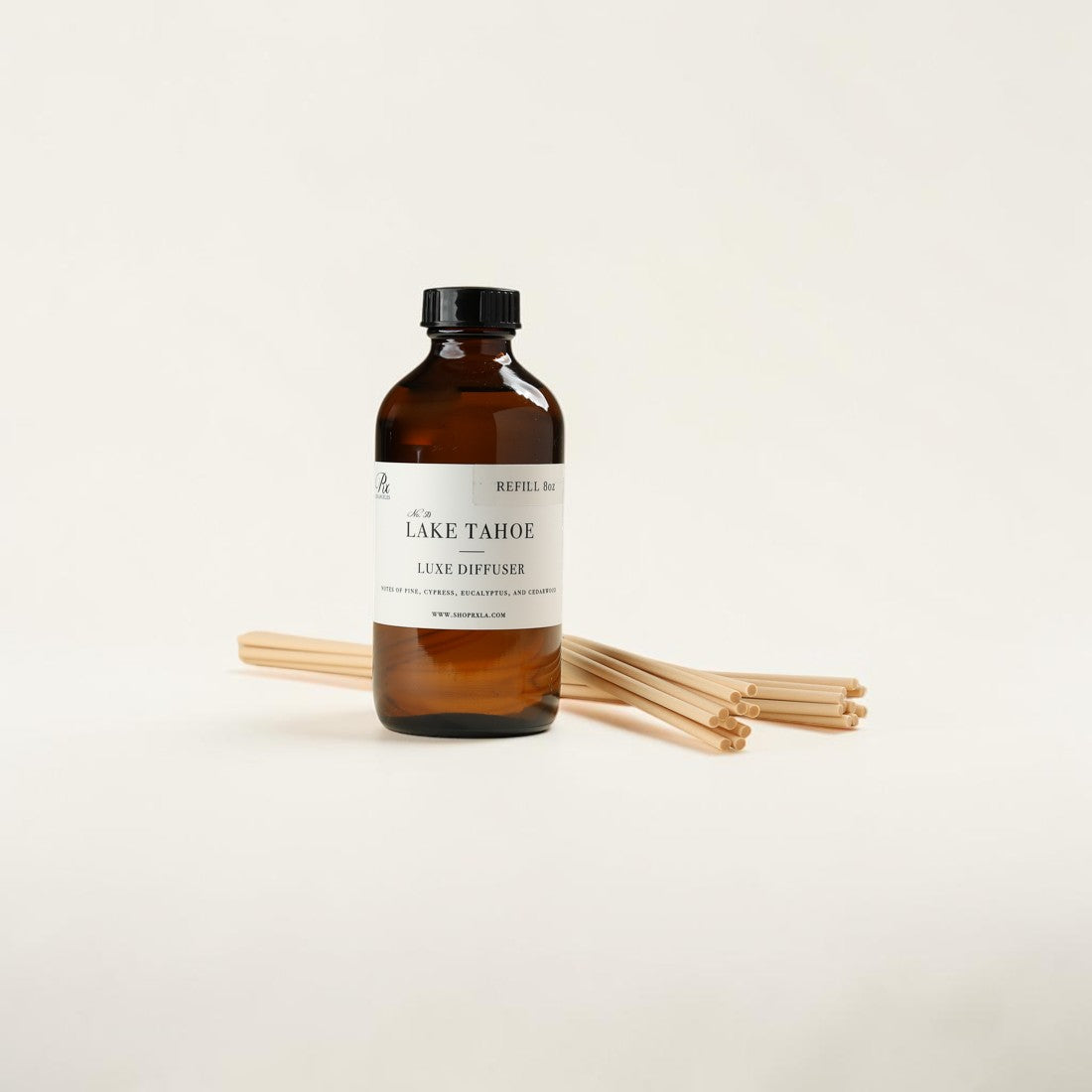An amber glass refill bottle labeled Lake Tahoe Luxe Diffuser with a black cap, standing upright on a white surface with several natural wood diffuser sticks placed behind it.