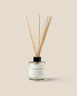 A reed diffuser bottle with fiber reeds, a clear glass container, and a black cap, labeled 'Lavender Oak', set against a solid beige background.