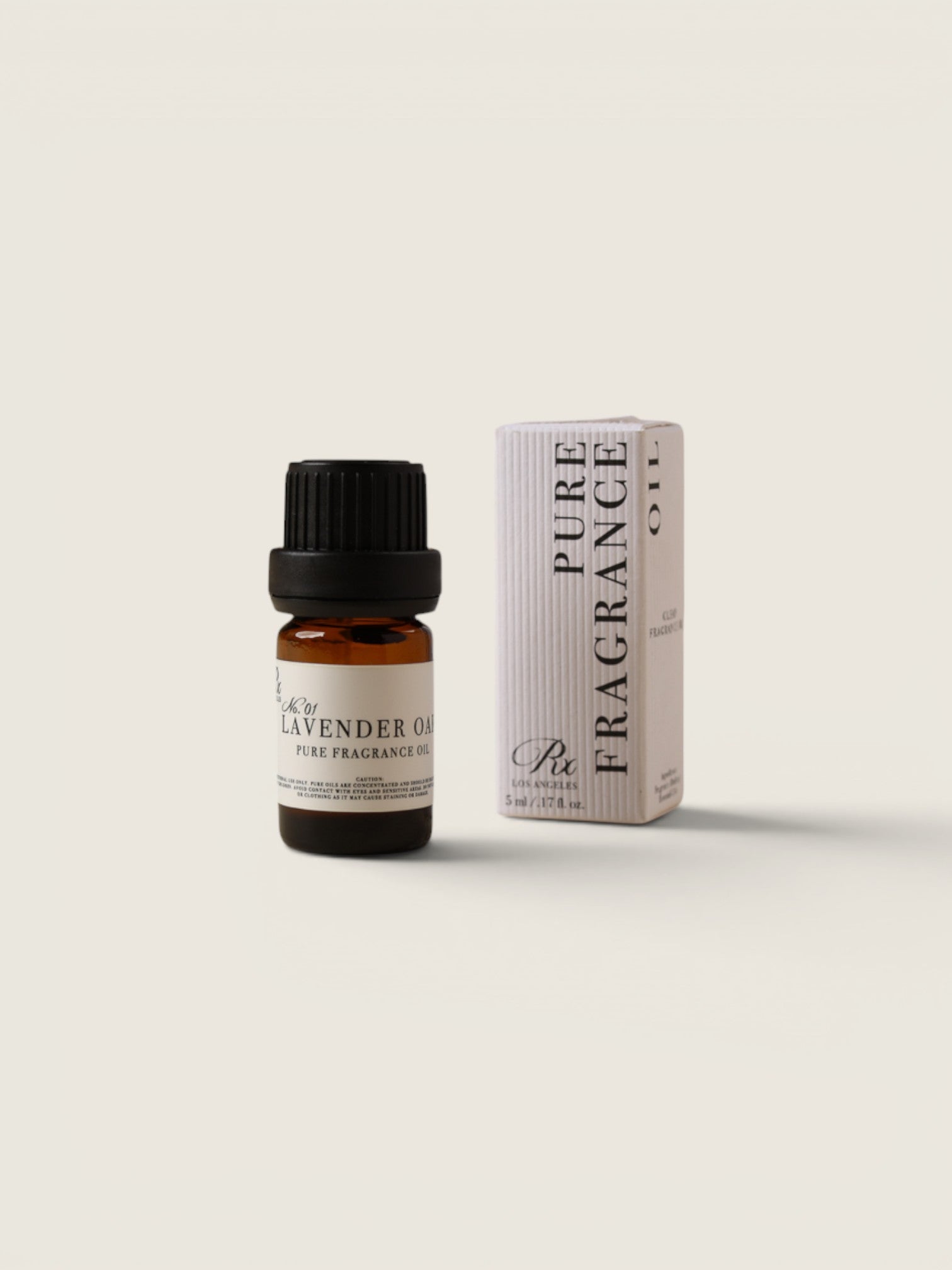 A small amber glass bottle of lavender oil with a black cap stands next to its white box packaging on a warm off-white background. The box is labeled 'PURE FRAGRANCE' and 'Lavender Oil'.