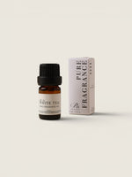 A small amber glass bottle of white tea pure fragrance oil with a black cap stands next to its white and black packaging box on a light beige background.