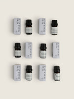 Nine small fragrance bottles with black caps arranged in a 3-by-3 grid alongside matching white rectangular boxes on a light beige background. The labels show Pure Fragrance oil