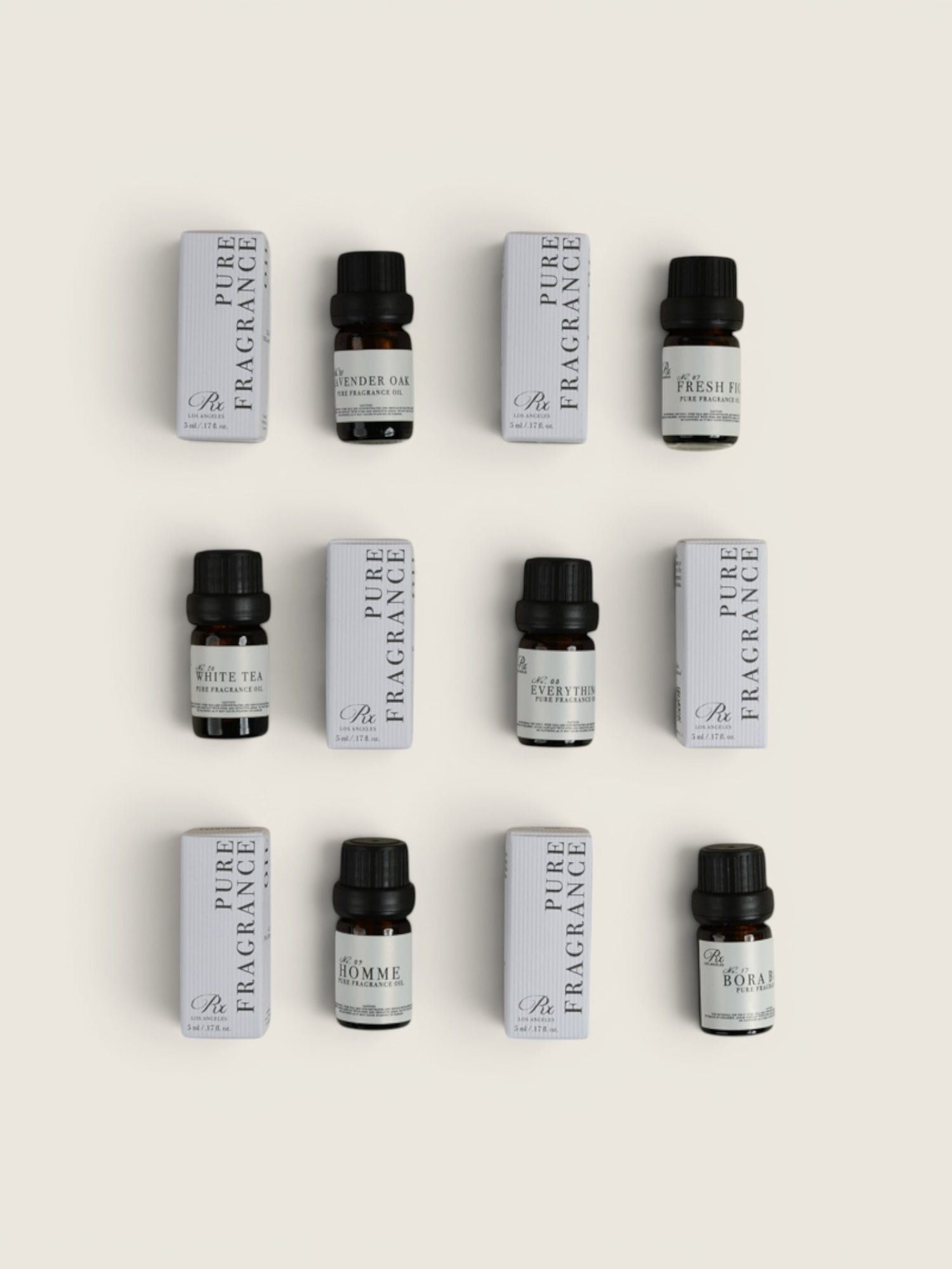 Nine small fragrance bottles with black caps arranged in a 3-by-3 grid alongside matching white rectangular boxes on a light beige background. The labels show Pure Fragrance oil