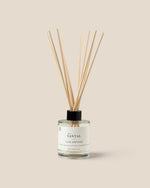 A Santal Luxe Diffuser with a clear glass bottle, white label, and natural wood reeds, set against a solid beige background.