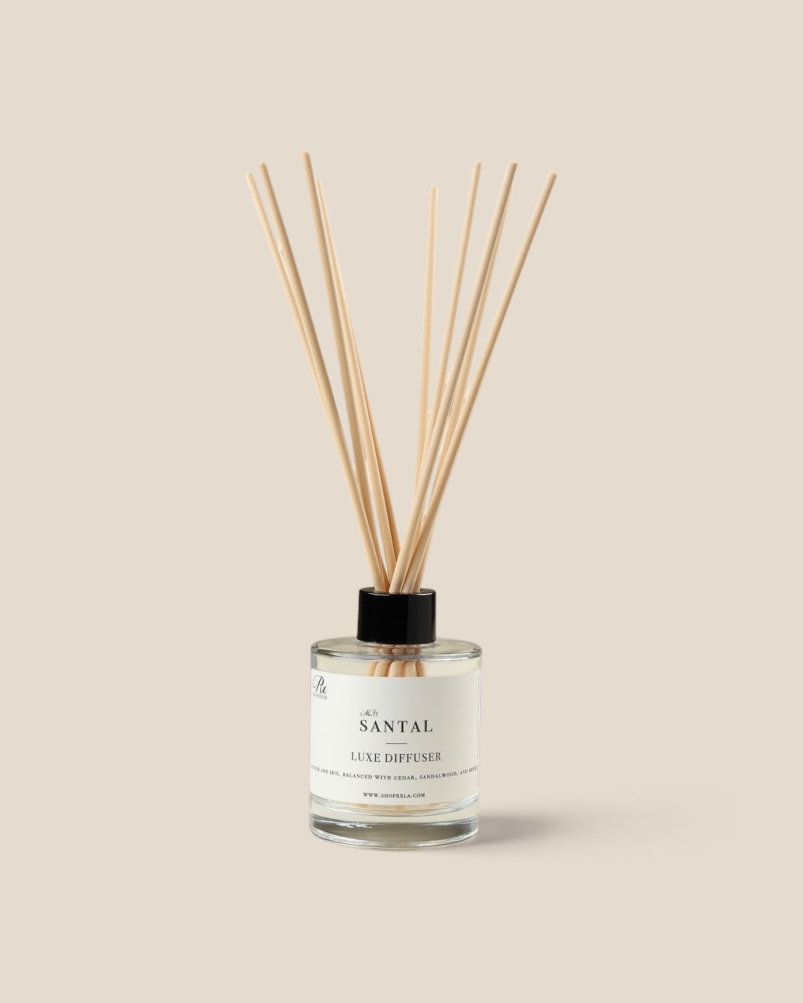 A Santal Luxe Diffuser with a clear glass bottle, white label, and natural wood reeds, set against a solid beige background.