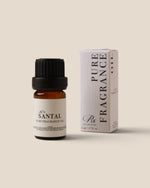 A small amber glass bottle of pure fragrance oil with a black cap stands next to its white box packaging on a solid beige background. The label reads 'Santal Pure Fragrance Oil' and the box reads 'PURE FRAGRANCE OIL' with 'Ric' branding.
