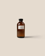 An amber glass bottle with a black cap and a white label reading 'SANTAL', centered on a solid beige background.