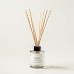 A reed diffuser bottle with a clear glass base, white label, black cap, and natural wood reeds, photographed on a clean off-white background. The label reads 'THE SANTAL' and 'REED DIFFUSER'.