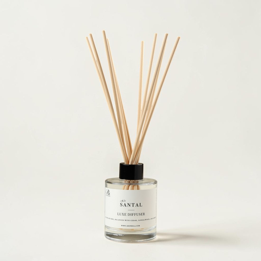 A reed diffuser bottle with a clear glass base, white label, black cap, and natural wood reeds, photographed on a clean off-white background. The label reads 'THE SANTAL' and 'REED DIFFUSER'.