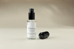 A clear glass spray bottle labeled 'SANTAL' with a black pump and a separate black cap placed beside it on a warm beige studio background.