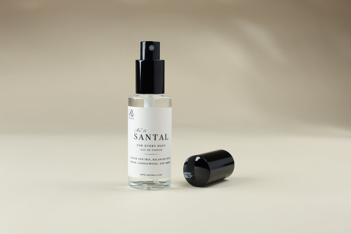 A clear glass spray bottle labeled 'SANTAL' with a black pump and a separate black cap placed beside it on a warm beige studio background.