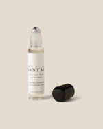 A clear glass rollerball perfume bottle with a white label and black text, shown next to its detached black cap on a solid beige background.