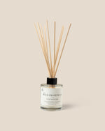 A reed diffuser bottle with natural wooden reeds, a clear glass container, and a black cap, labeled 'The Wild Grapefruit' by 'RXLA', set against a solid beige background.