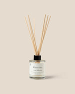 A reed diffuser bottle with a clear glass body, white label reading 'WHITE TEA', and fiber reeds extending upward against a plain warm beige background.